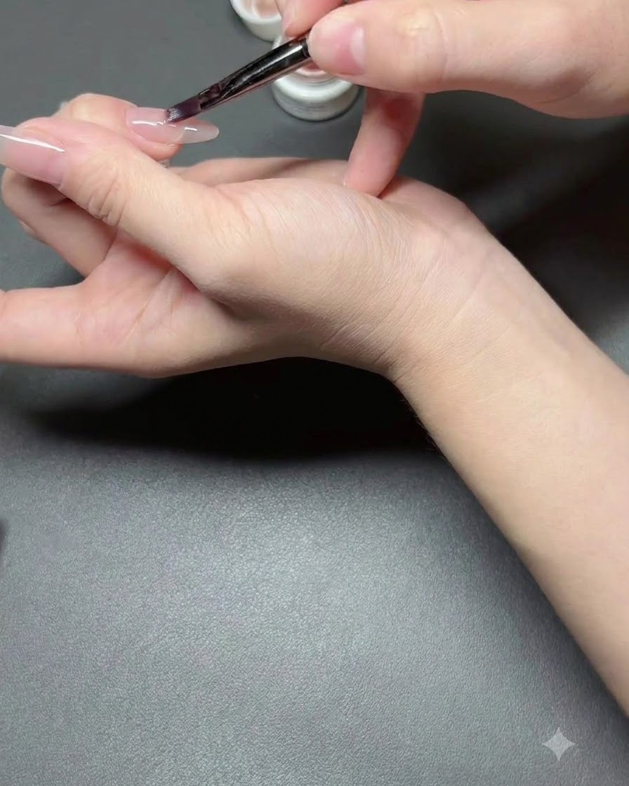 A nail brush applying a thin coat of nude semi-sheer polish onto a fingernail.