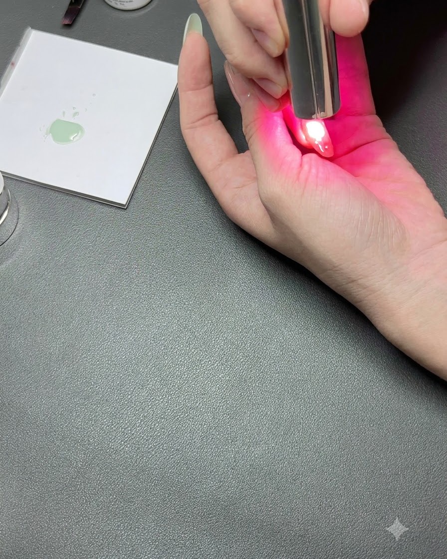 A hand under a compact UV nail lamp with a pink light curing a detailed nail.