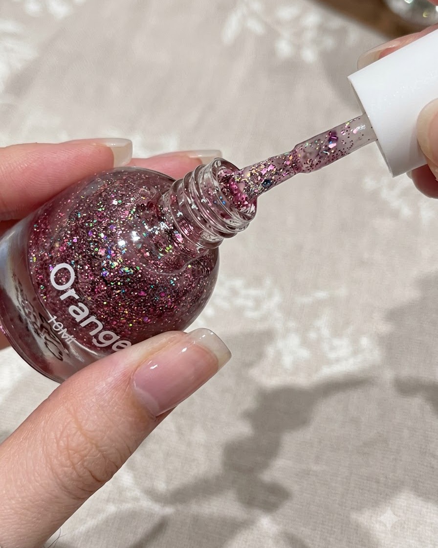 A brush applying a thin layer of pink glitter gel onto the upper half of a natural fingernail.