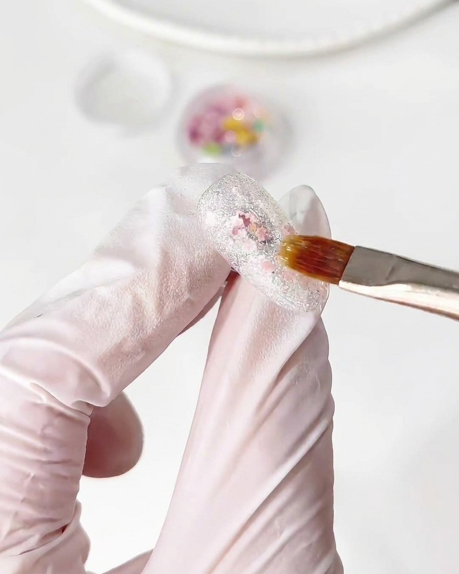 A brush placing small colorful flower glitter pieces onto a wet base gel layer on a nail.