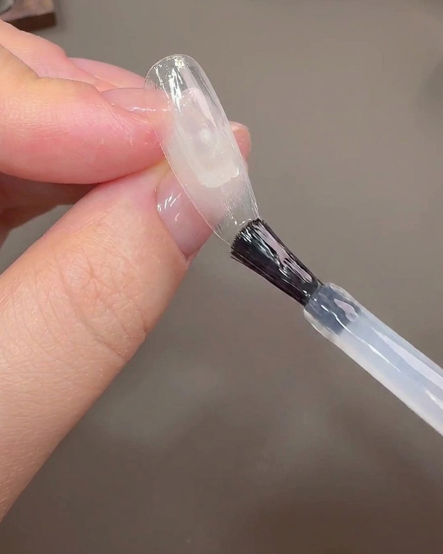 A nail brush applying builder gel onto a clear nail tip to cover the growth line.