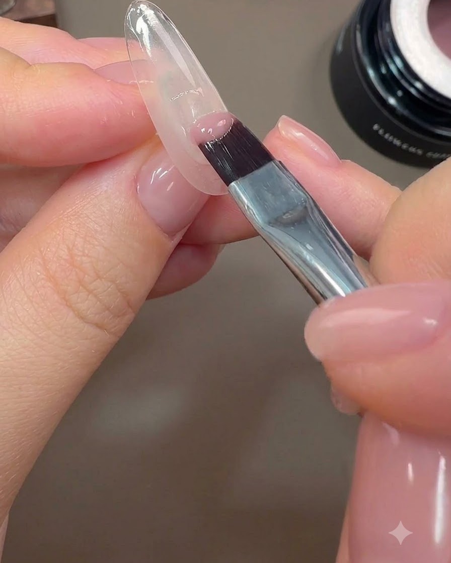 A nail brush applying a half-cover of nude pink color gel to the posterior edge of a nail.