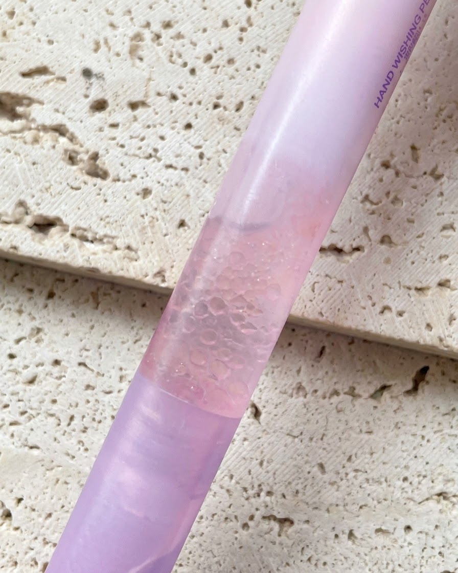 A pink gradient pearl essence cuticle oil pen held against a textured background.