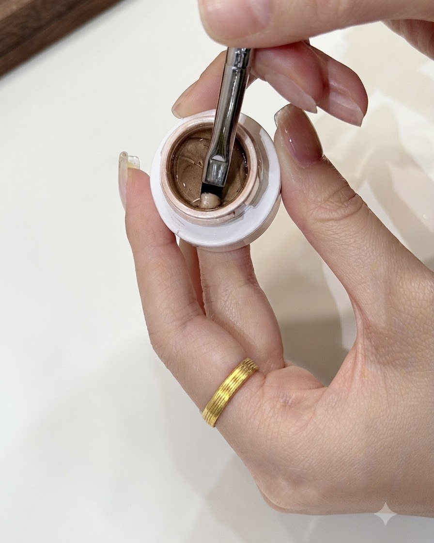 A nail art brush scooping a small, mung bean-sized portion of champagne gold gel from an open jar.
