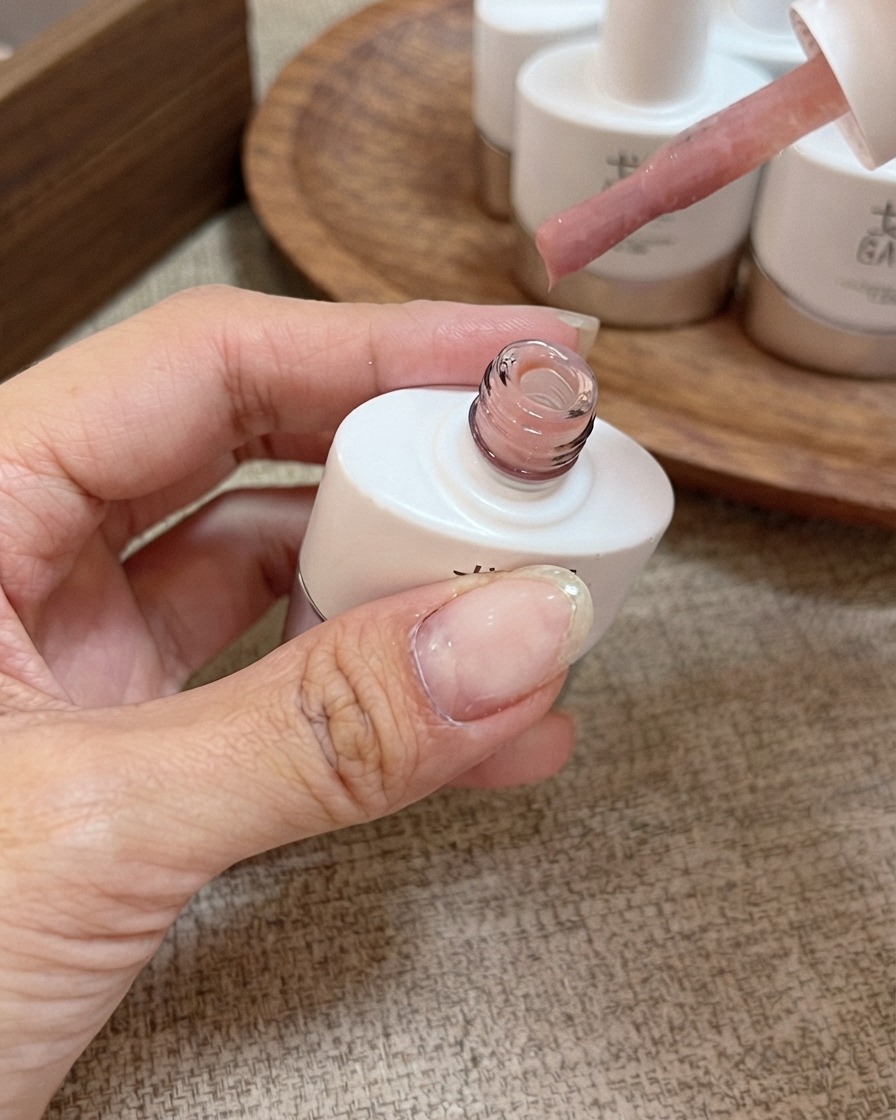 A brush applicator with honey peach pink nude gel polish held above an open bottle.