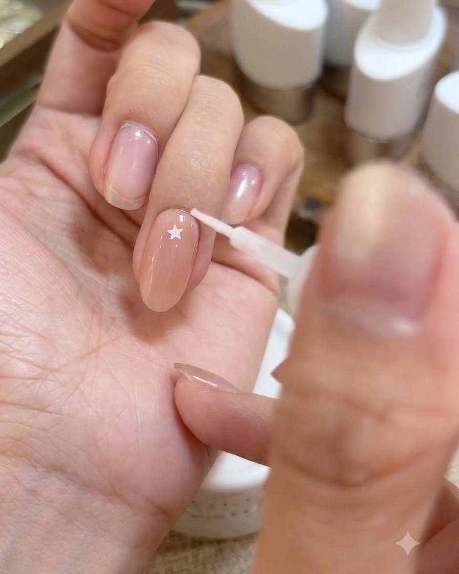 A tool placing a small iridescent star sticker onto the glossy base gel surface of a fingernail.