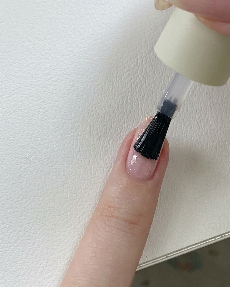 A brush applying a clear, glossy top coat over a pink glitter gel nail.