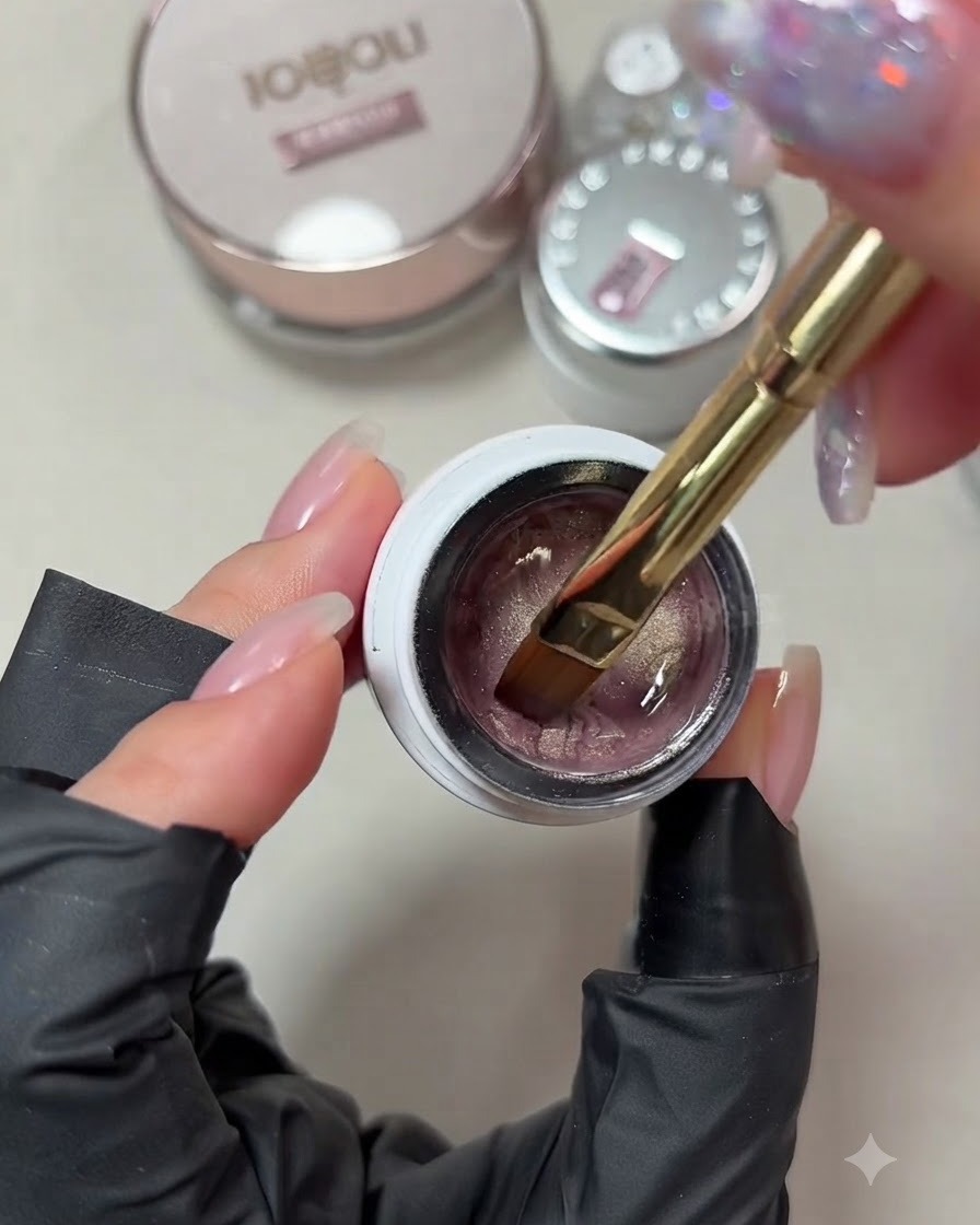 A close-up view of a gel brush dipping into a jar of pale pink champagne cat-eye gel polish.