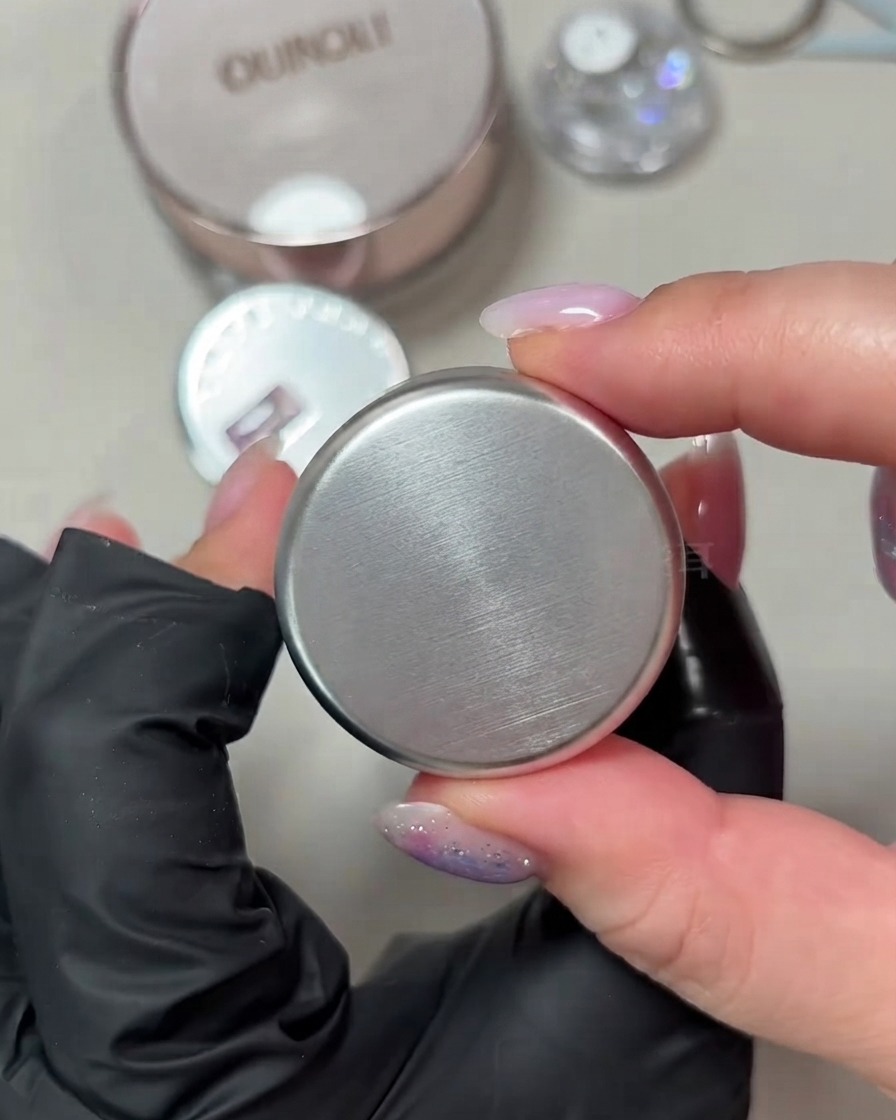 A hand holding up a small round jar of porcelain white shimmer cat-eye gel against a neutral background.