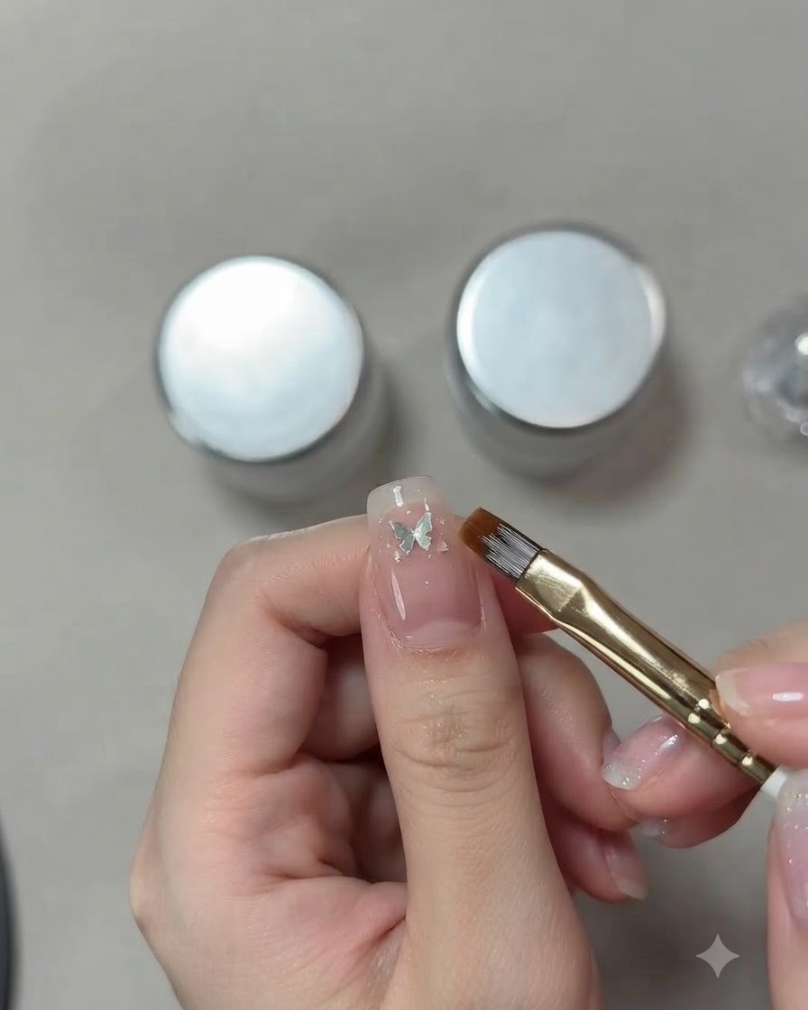 A detailed close-up of a nail artists hand using a fine brush to position a small silver butterfly sequin on a pink cat-eye nail.