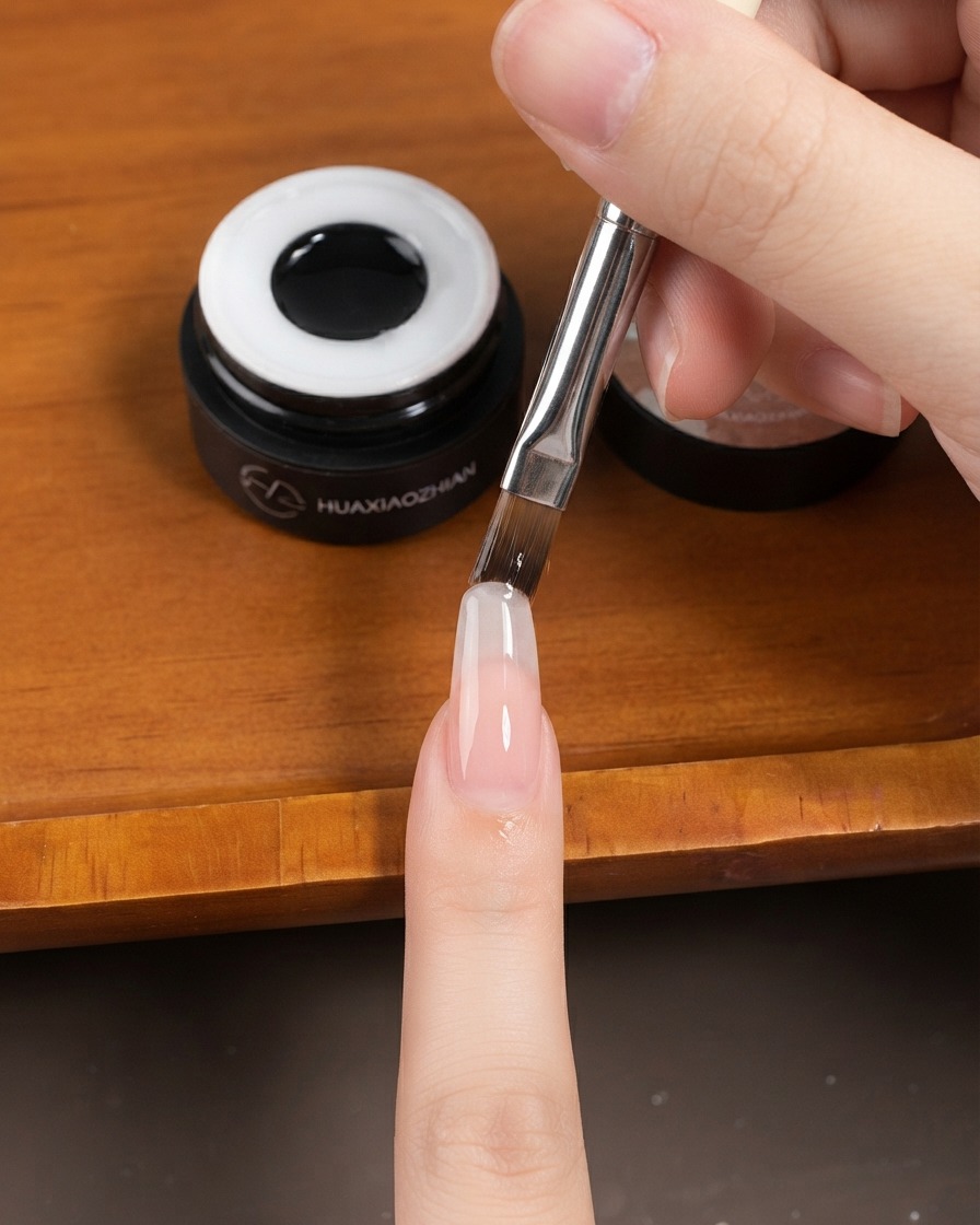A flat gel brush applying a sheer layer of gel onto a long almond-shaped fingernail.