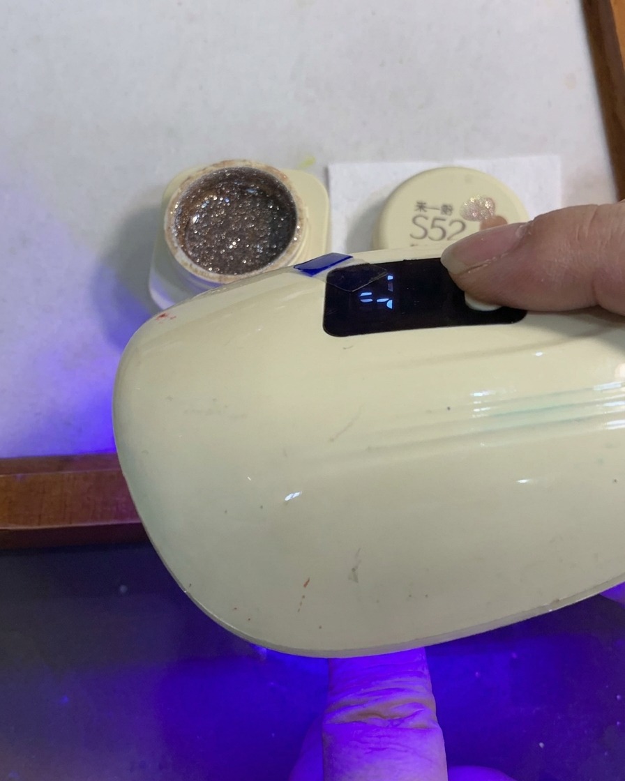 A finger with a glitter gradient nail curing under a small portable UV/LED nail lamp.