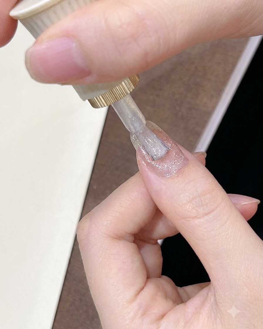 A brush applying a sheer, glowing pinkish-brown gel polish across the surface of a manicured nail.