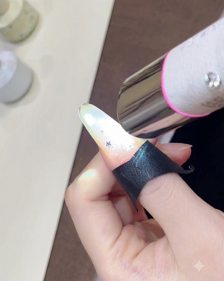 A hand holding a mini UV lamp over a nail decorated with a sparkling glitter gradient and small star sequins.