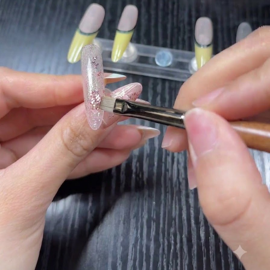 A close-up of a brush applying pink and silver holographic glitter flakes over a sheer base on a practice nail tip.