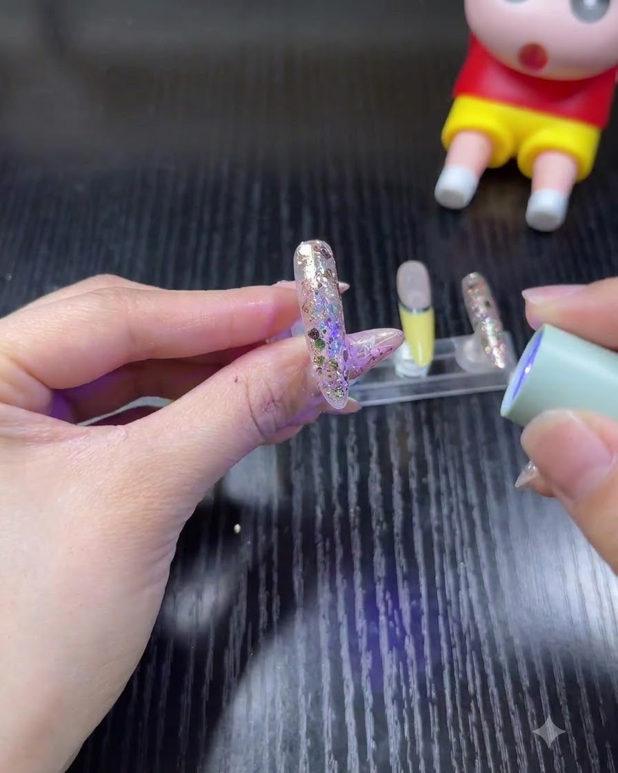A hand holding a mini, handheld UV/LED lamp over a glittery nail to cure the gel.