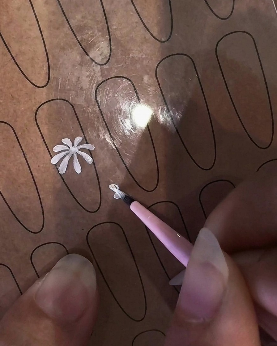 A close-up of a hand holding a fine liner brush to paint a white daisy petal onto a clear nail practice sheet.