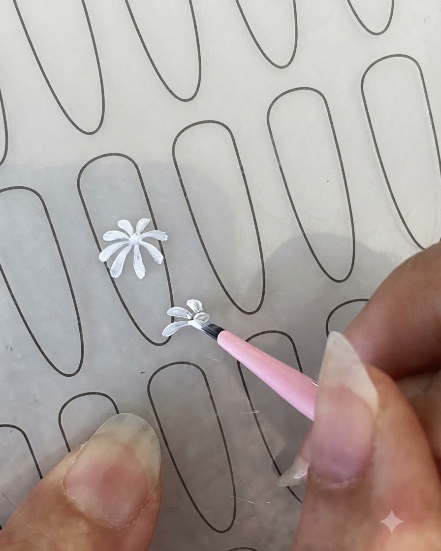 A finished white daisy petal outline on a practice nail sheet next to a fine liner brush.