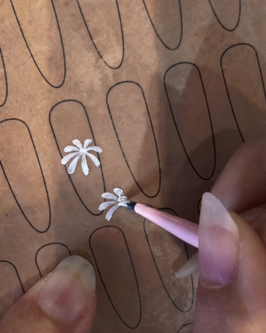 A fine liner brush beginning a second smaller white petal on a practice nail sheet.
