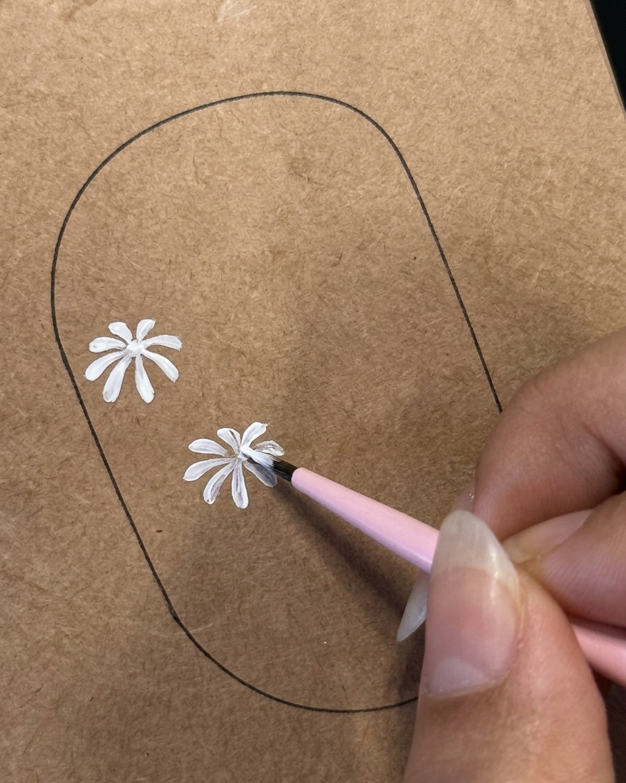 A fine liner brush detailing white petals on a second daisy design painted on a brown nail practice template.