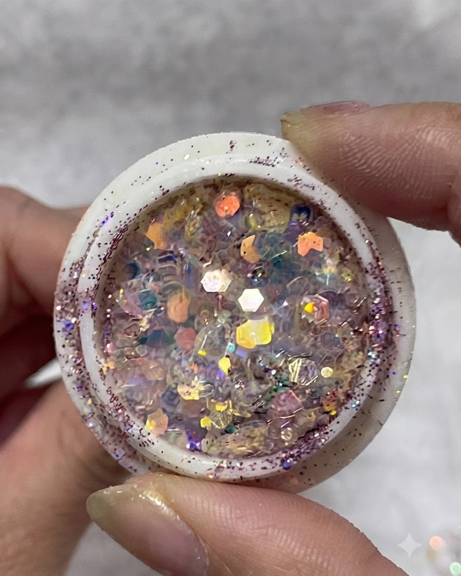 A jar filled with multi-colored iridescent glitter flakes, sequins, and small stars held between fingers.