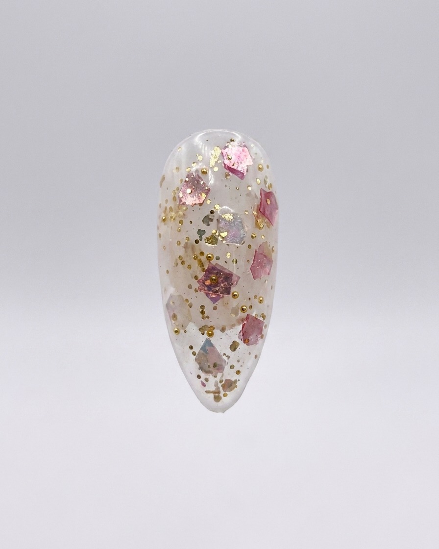 A clear press-on nail featuring embedded pink and gold glitter flakes, shown isolated on a light gray background.