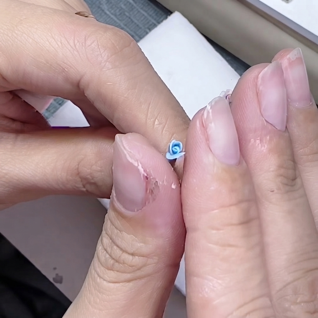 Fingers delicately pinching the base of a small blue petal around a sculpted rose core.