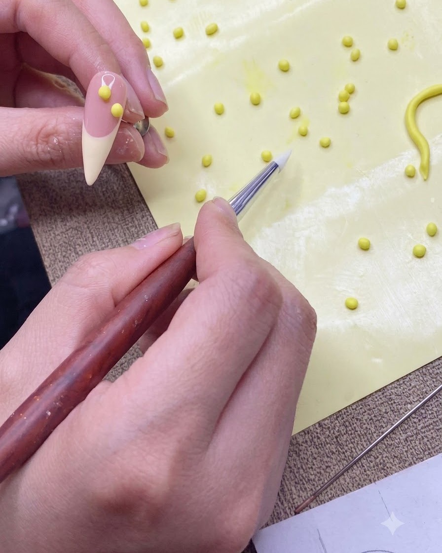 A silicone shaping pen transferring a small yellow gel ball onto a nude-colored practice nail tip.