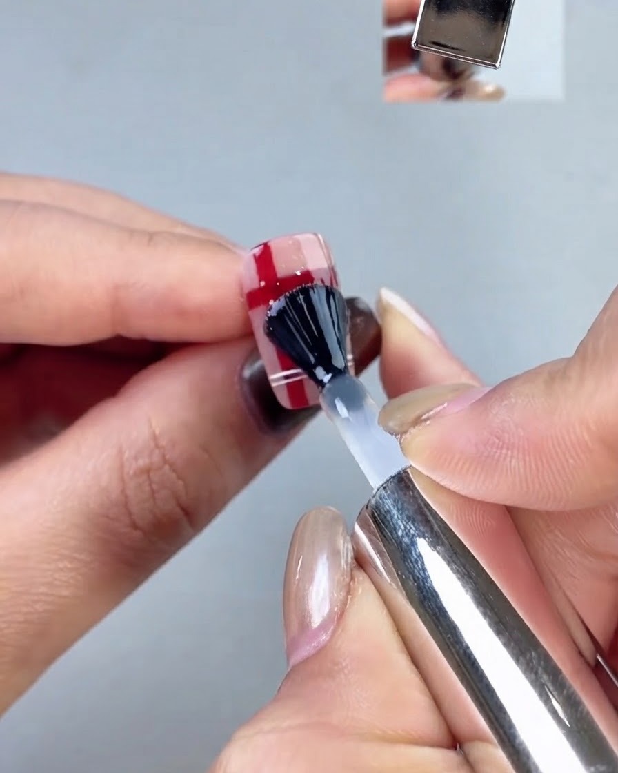 A black-capped nail brush applying a clear top coat layer over a plaid nail design.