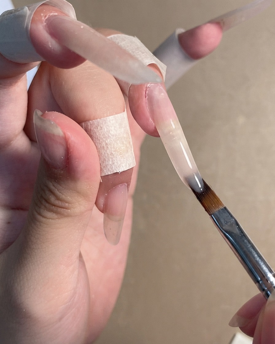 A nail brush applying a sheer nude gel polish onto a long stiletto nail extension.