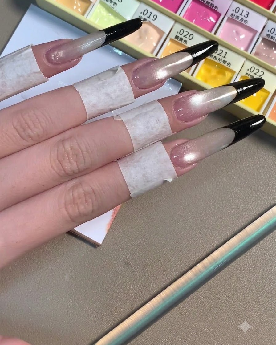 Four long stiletto nails displaying completed deep black French tips over a shimmering grey-toned base.