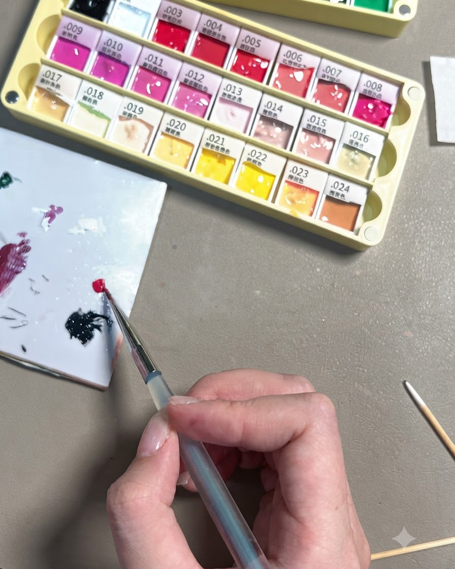 A hand using a fine detail brush to deposit a small dot of bright rose red gel onto a white ceramic mixing palette.