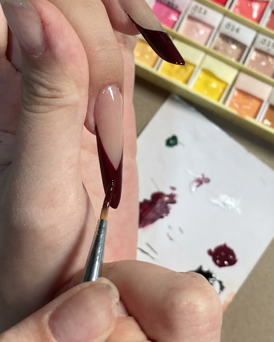 A fine brush adding small dark plum accents and petal shapes onto a black French tip.
