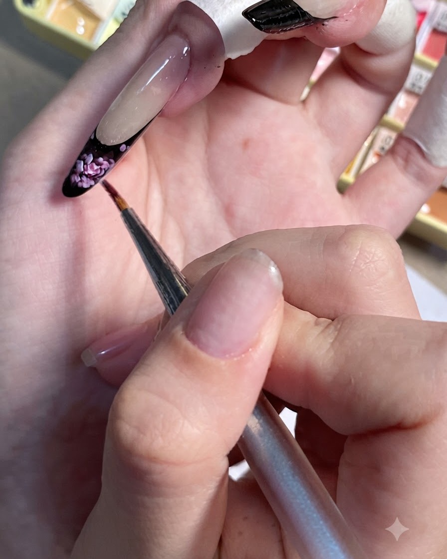 A detailing brush dotting small light pink accents onto the black French tip of a painted nail.