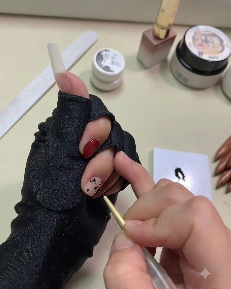 A close-up of a hand using a fine golden liner brush to pull black gel into leaf shapes from dots on a nude base nail.