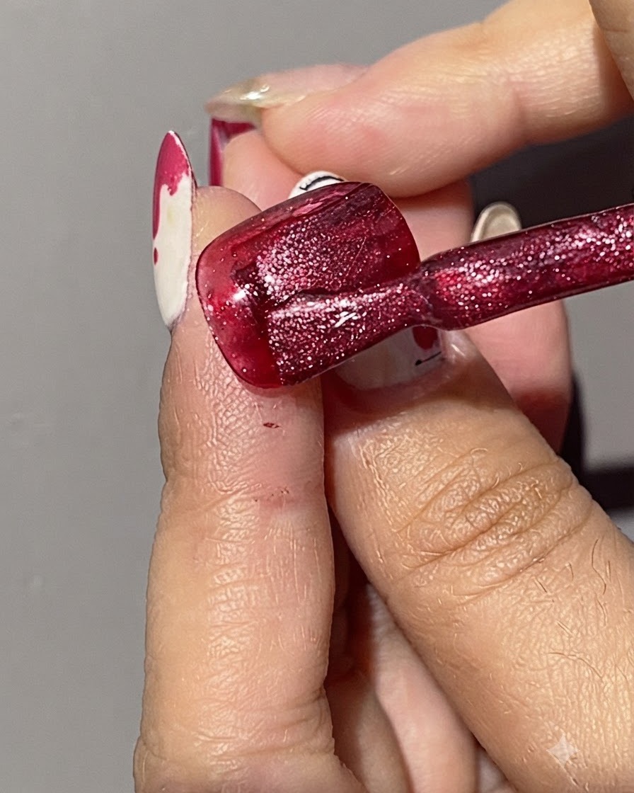 A brush applying a second coat of shimmering red cat eye gel polish over an already red-painted nail.
