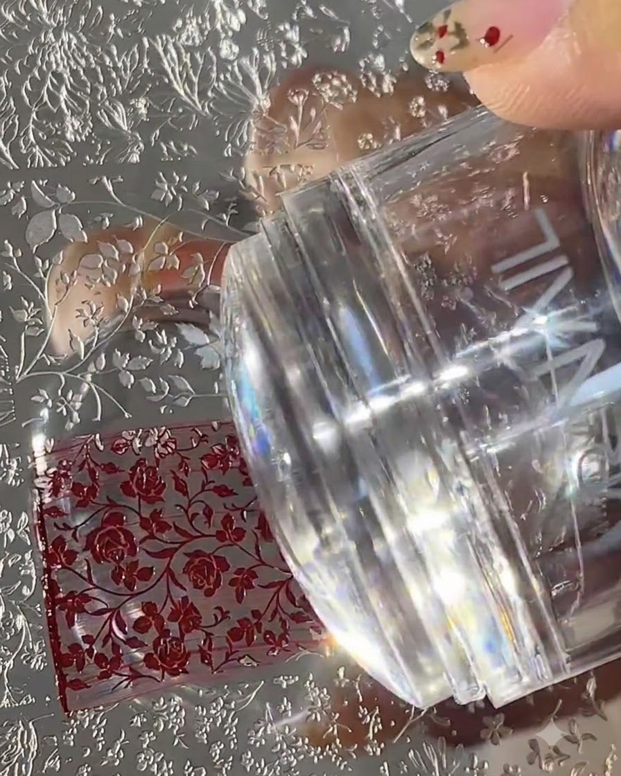 A clear silicone nail stamper pressing down onto an engraved metal stamping plate filled with red floral patterns.