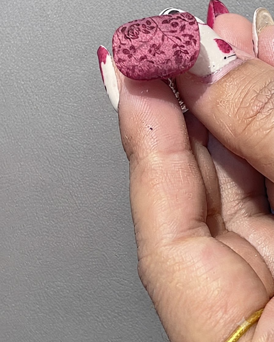 A manicured nail with a stamped red floral pattern being lightly coated with white crystal powder using a spatula.