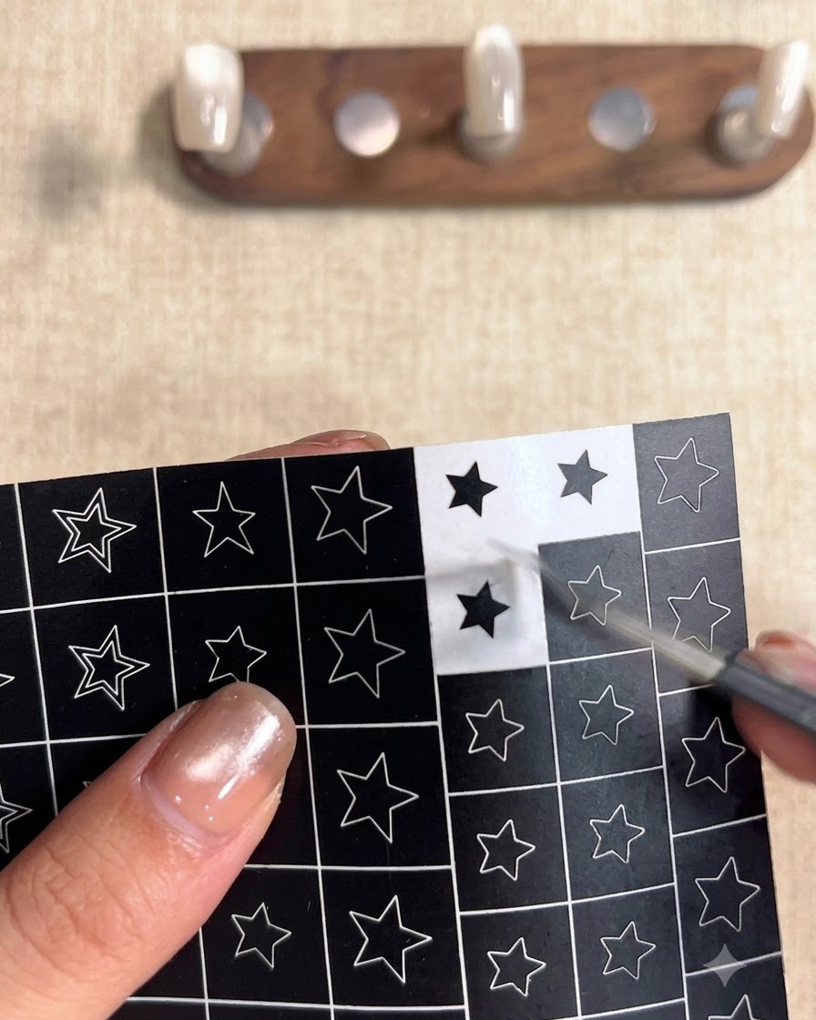 A hand using tweezers to peel a black square sticker with a hollow star cutout from a sheet of star designs.