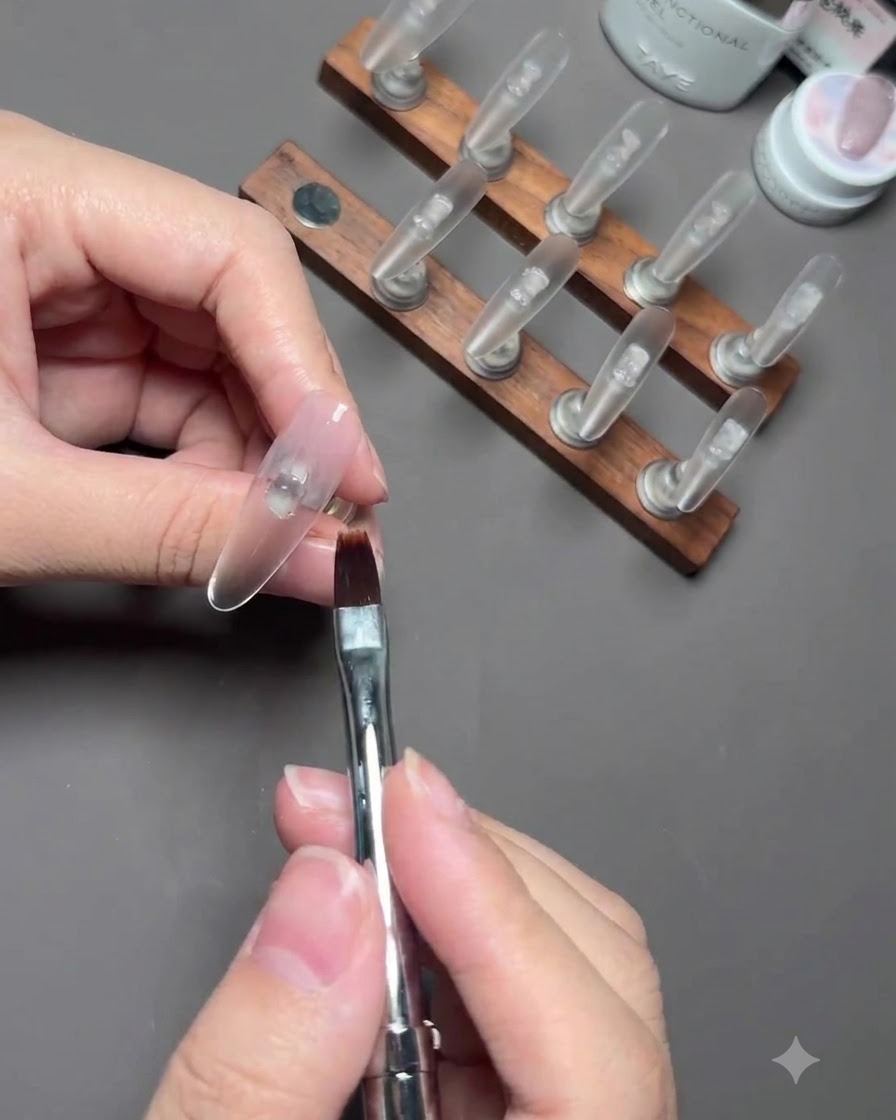 A nail brush blending sheer nude gel polish on a thumb nail mounted on a stand.