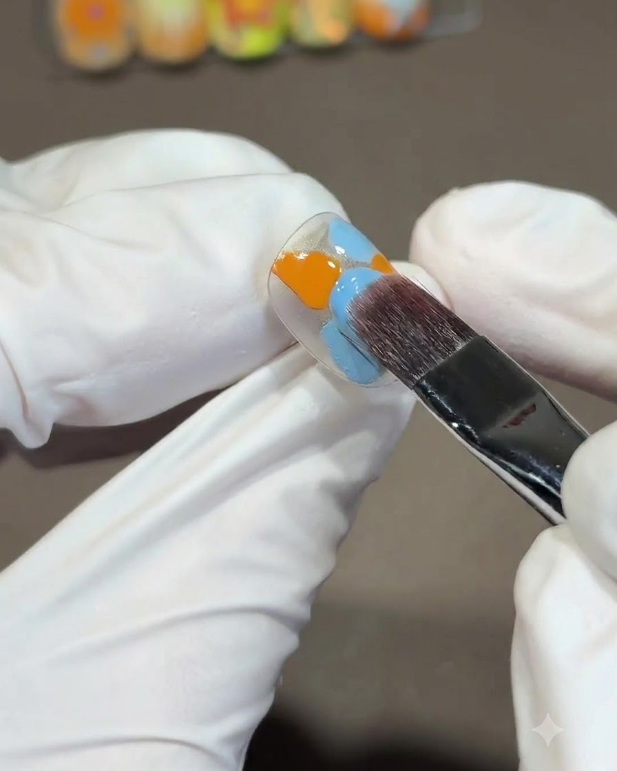 A large square brush blending orange and blue gel polishes on a nail tip using an S-shaped downward stroke.