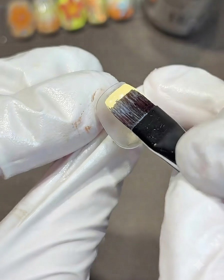 A gloved hand using a flat gel brush to paint a clear practice nail with solid yellow gel polish.
