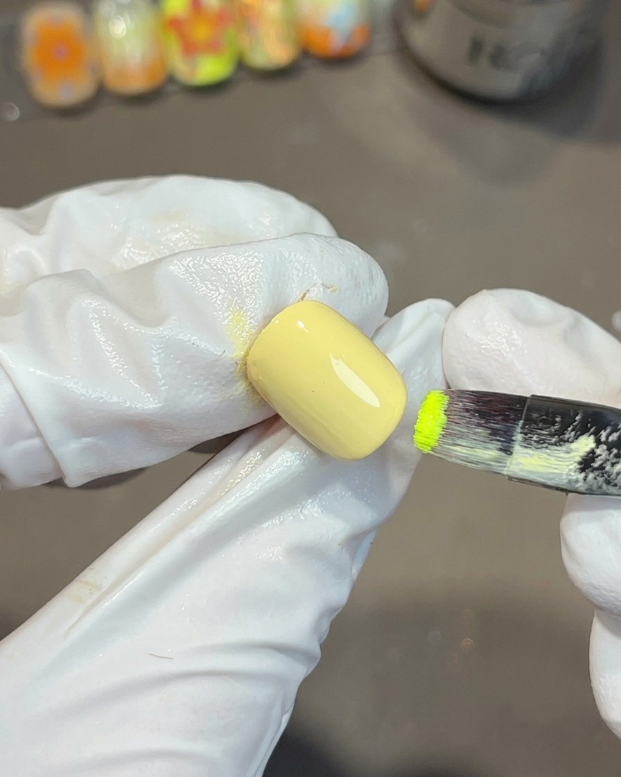 A gloved hand using a flat nail brush to apply vibrant green gel polish onto the corner of a yellow painted nail.