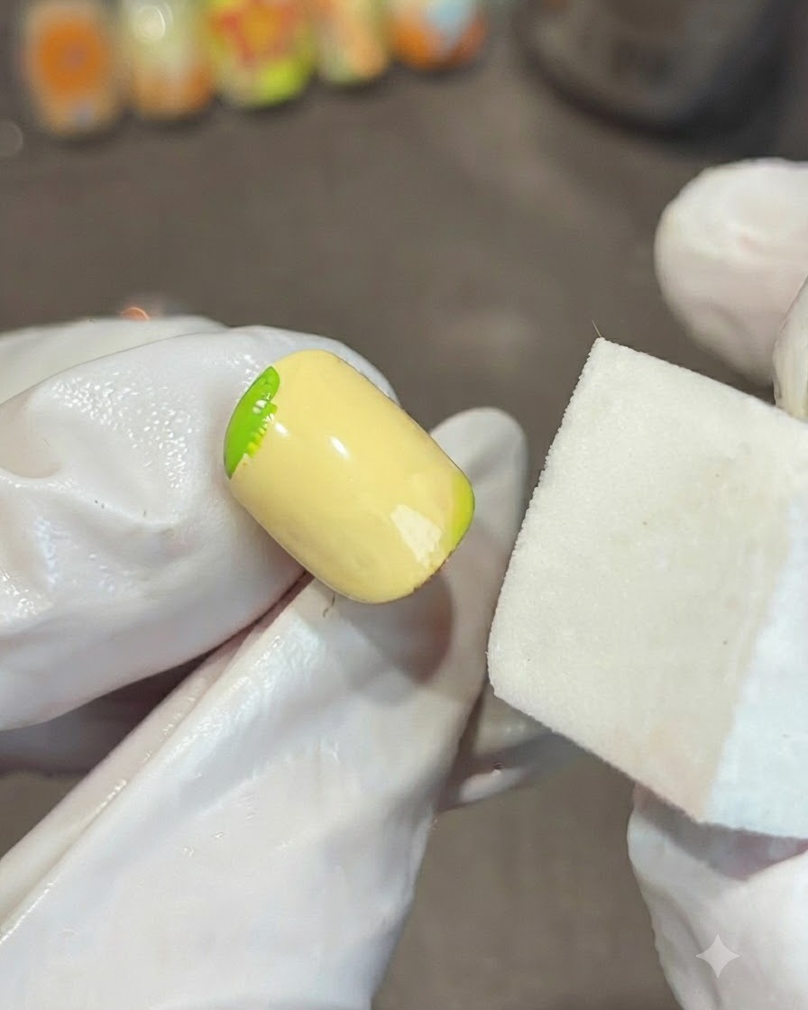 A gloved hand holding a white sponge block next to a yellow nail, blending a green gel gradient on the corner.