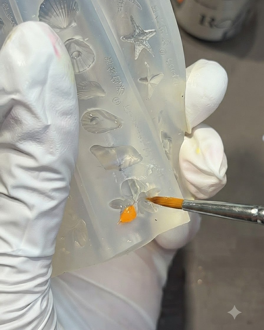 A gloved hand using a detail brush to fill a lily flower shape inside a clear silicone mold with bright orange gel.