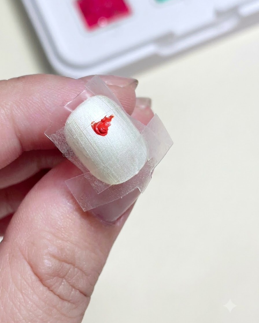 A close-up of a prepped nail with a clear taped square in the center, featuring a bright dab of red gel paint.