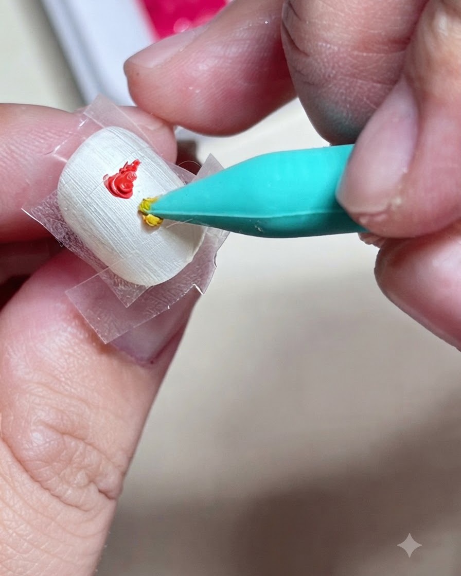 A close-up of a silicone nail tool applying a drop of yellow gel paint directly below a red dab inside a taped square on a nail.