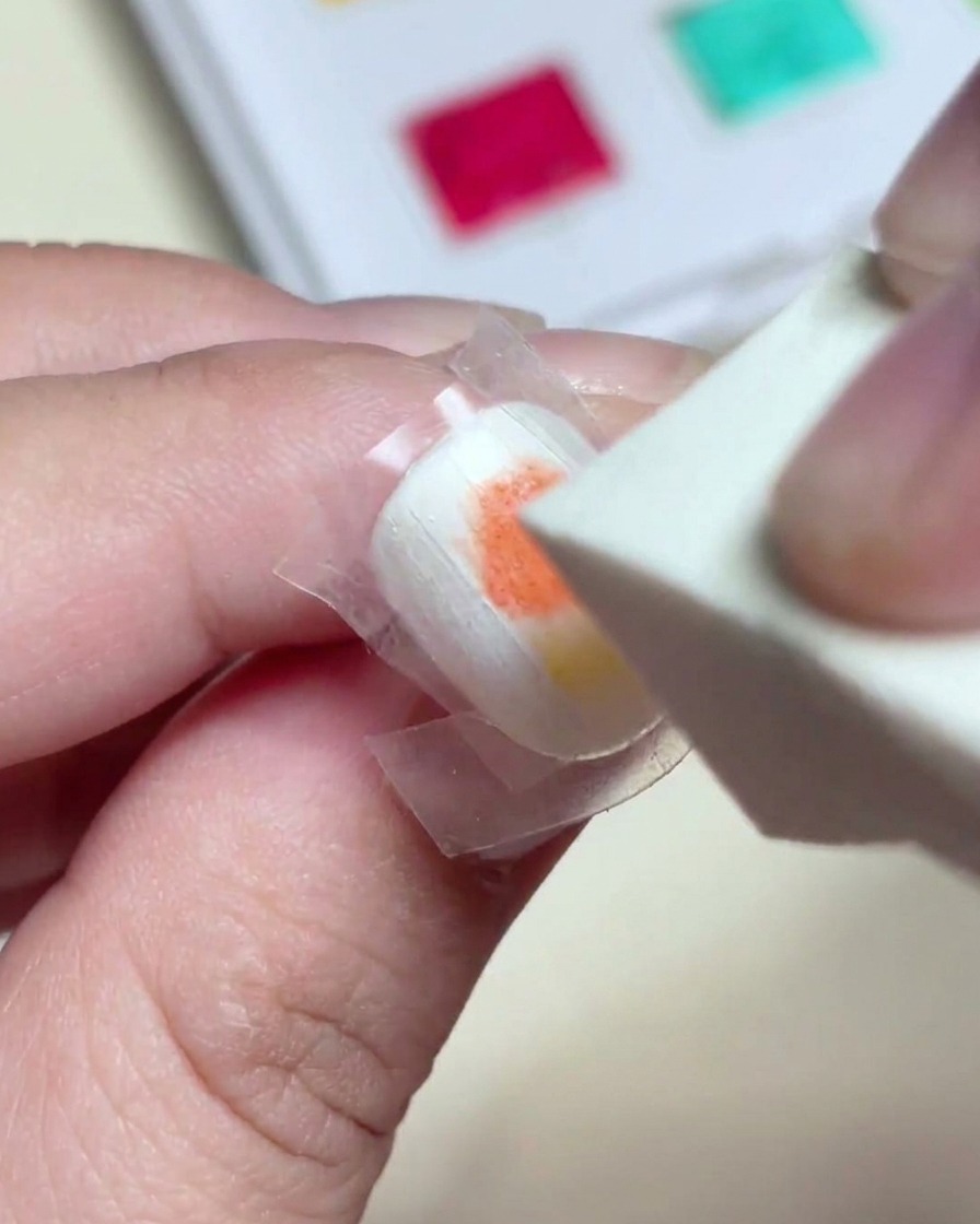 A makeup sponge dabbing gently over wet red and yellow gel paint to create a seamless gradient on the nail.