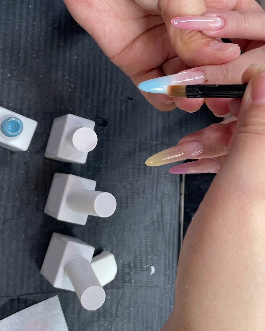 An ombre blending brush being held flat against a fingernail to blend a light blue gel gradient downward.