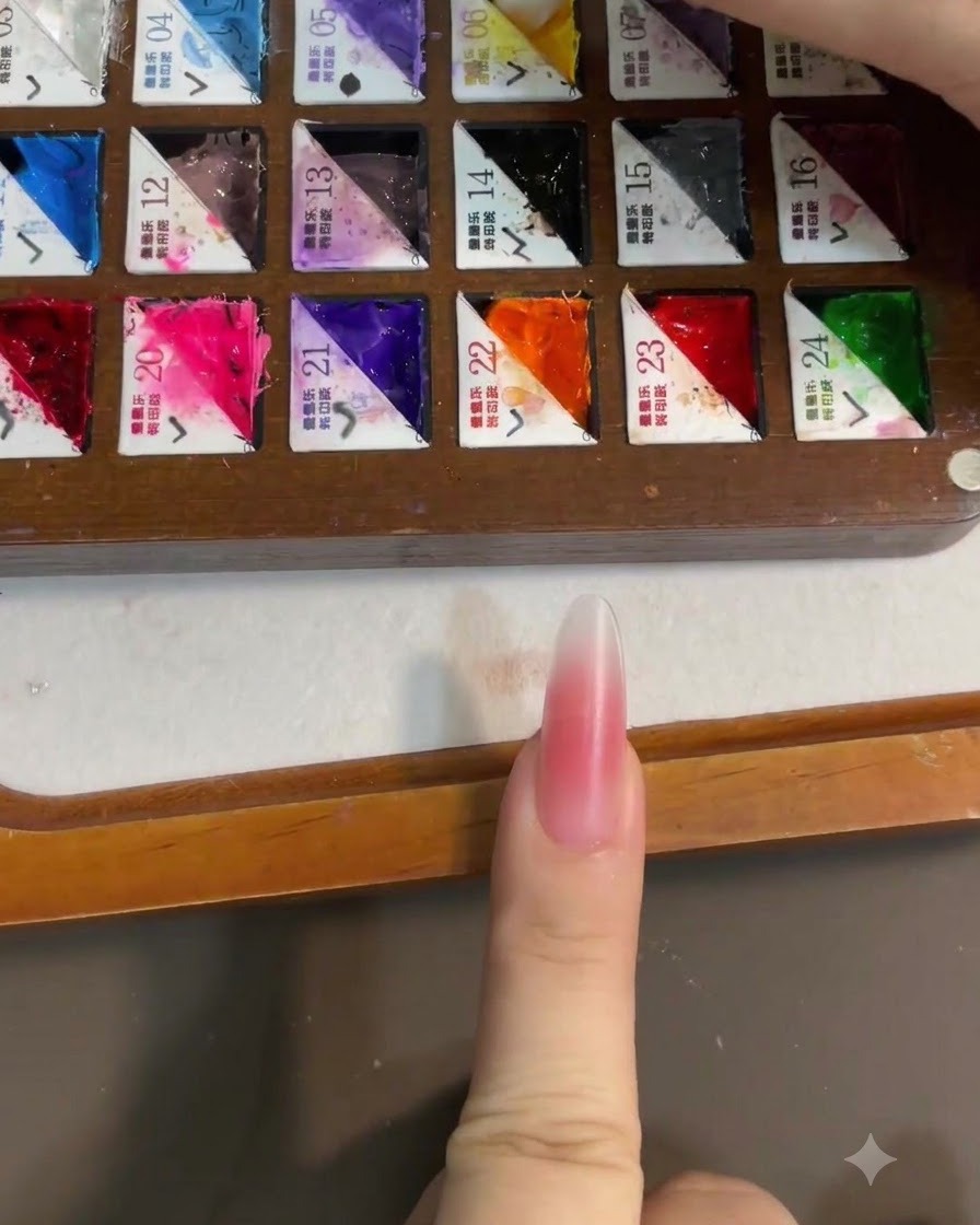 A close-up of a brush picking up bright yellow gel polish from a partitioned painters palette, with a pink blush nail in the foreground.